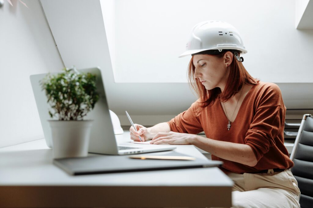pexels-photo-5583254-5583254 A focused female architect in a hard hat writing notes while working on a laptop indoors.